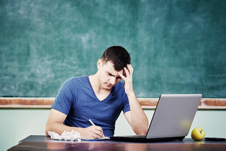Sad upset overworked man studying in front of  stack of books with a blackboard behind him.の写真素材
