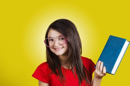 Smiling Portrait of a cute little schoolgirl loving to learn, holding with hands a book and wearing...