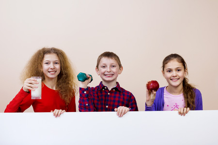 Happy smiling group of kids, friends, boys and girls, holding milk,dumbbell, red apple showing white blank placard, board,poster.の写真素材