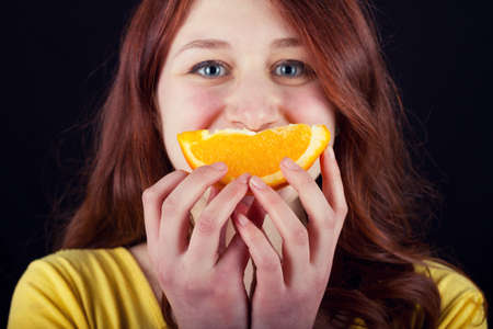 Young woman smiling and eating a fresh slice of orange over black background.の写真素材