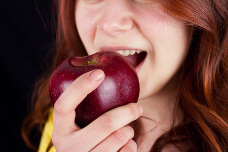 Young woman smiling and eating a fresh red apple over black background.