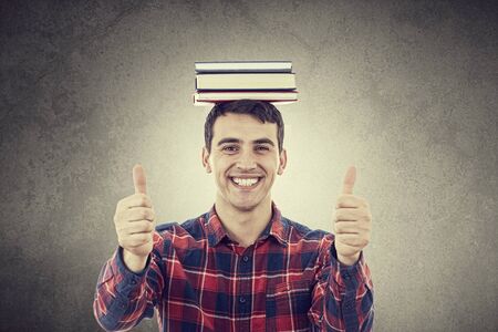 Smiling excited young man with thumb up holding a stack of books on his head isolated on white background.Close-up shot of Happy smiling student with thumb up in shirt over white background.の写真素材