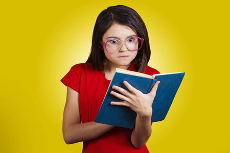 Surprised, Amazed cute little schoolgirl with a open book on her head, isolated over yellow background.の写真素材