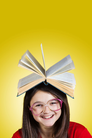 Smiling Portrait of a cute little schoolgirl loving to learn with a book on her head, isolated over yellow background.の写真素材