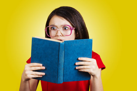 Surprised, Amazed cute little schoolgirl with a open book on her head, isolated over yellow background.の写真素材