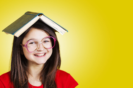 Smiling Portrait of a cute little schoolgirl loving to learn with a book on her head, isolated over yellow background.の写真素材