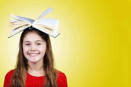 Smiling Portrait of a cute little schoolgirl loving to learn pointing up with a open book on her head isolated over yellow background.の写真素材