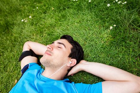 Sport fitness man relaxing listening to music after training outdoor in a city park . Young male athlete resting relaxing lying on grass after running and training exercise outside in summer.の写真素材