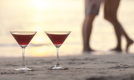 Two cocktails on the beach with couple kissing in the background, concept about romantic honeymoon in tropicsの写真素材