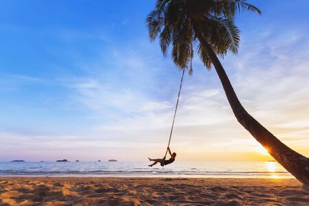 Young happy woman relaxing on a swing attached to a palm tree on a paradise beach at sunset while on vacation in a tropical countryの写真素材