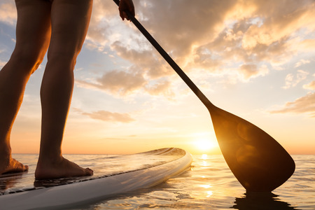 Stand up paddle boarding on a quiet sea with warm summer sunset colors, close-up of legsの写真素材