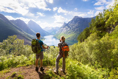 Two hikers at viewpoint in the mountains enjoying beautiful view of the valley with a lake and sunny warm weather in summer, green trees aroundの写真素材