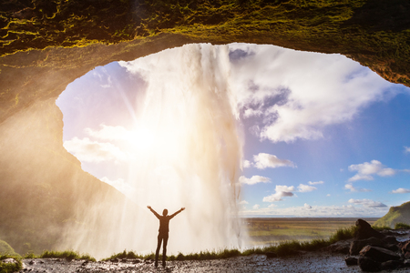 Happy female traveler feeling the power of Seljalandsfoss waterfall in the South of Iceland, person standing behind the stream, sunset light, intense icelandic experienceの写真素材