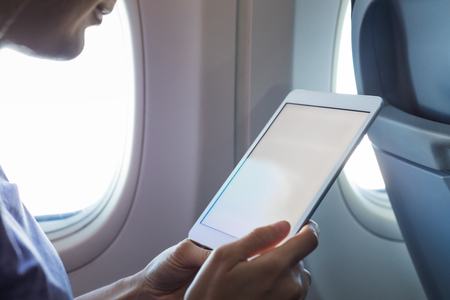 Passenger using tablet computer in airplane cabin during flight with wireless internet to read emails or an ebook, hands holding device with white empty screen for copy-space, onboard entertainmentの写真素材
