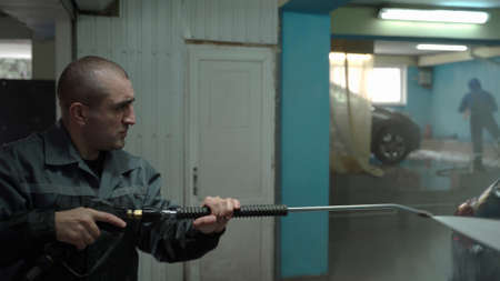 A Caucasian man works in a car wash service. A man in a workers uniform holds a water gun and washes...
