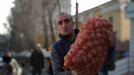Close-up portrait of a Caucasian nationality male loader worker holding a large bag of onions in his...