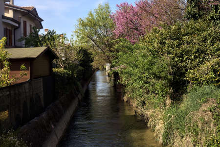 Stream of water bordered by houses and trees with reflections casted in the water on a sunny day in the italian countrysideの写真素材