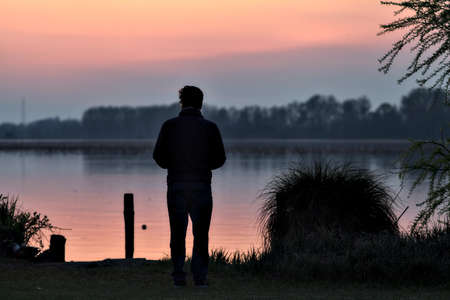 Man standing by the shore of a lake at sunsetの写真素材