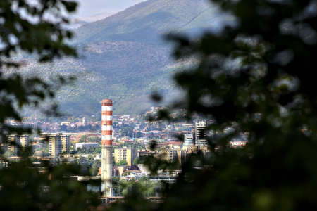 Red and white smokestack in a city seen from the distance framed by trees on a sunny dayの写真素材