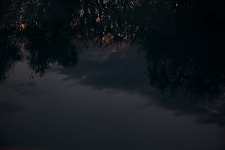 Mountain ridge with lights in the distance framed by trees at sunset casted in the waterの写真素材