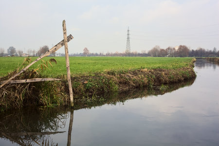 Stream of water in the countryside and a mooring pole by the shore with its reflection casted in the water on a cloudy dayの写真素材