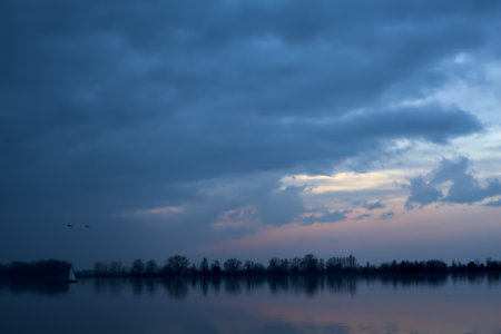 Lake at sunset on a cloudy day with a sailing ship in the distanceの写真素材