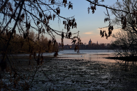 Skyline of a town seen from the distance over a lake at sunset framed by treesの写真素材