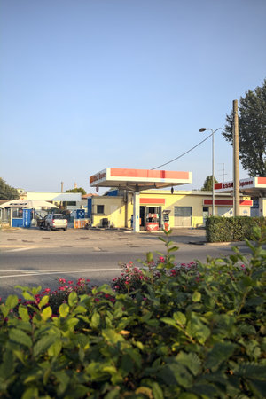 Msntova, Italy - October 2023 - Esso petrol station on a country road with a village and a chimney in the distance seen from behind a bush at sunsetのeditorial素材