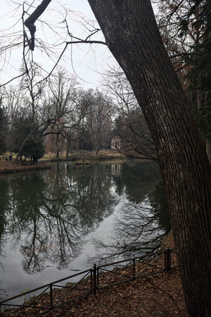 Pond in a park with a neoclassical temple on its shore and reflections of trees on a cloudy day in...
