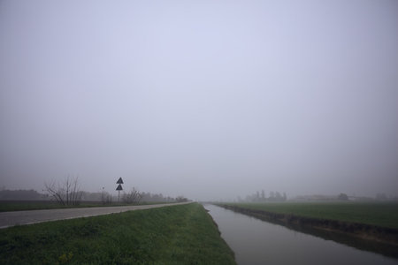 Country road next to a field bordered by a stream of water on a foggy day in autumnの写真素材