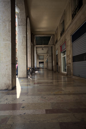 Tall and narrow porch under a building with closed shops and bicycle leaning on the columnsの写真素材