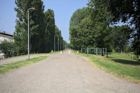 Paved trail on an embankment bordered by poplars in a park on a sunny day with a house next to its...