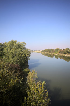 River bordered by trees on an embankment reflected in the water on a sunny day in the Italian...