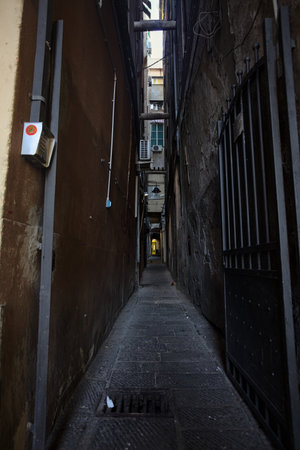 Extremely narrow alley in the dark with an open gate and an arch on the other end of it bordered by tall historical buildingsの写真素材