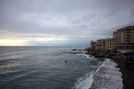 A row of houses on the top of a small cliff by the sea, and a boathouse nearby, on a cloudy day at sunset with waves crushing on a beach and on a breakwater that stretches to the seaの写真素材