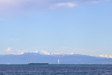 Cargo ships and a sail boat on a windy and sunny day with a mountain ridge covered by snow in the...