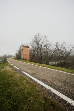 Road on an embankment, between a field and a grove, with a field below a side of the road, a brick...