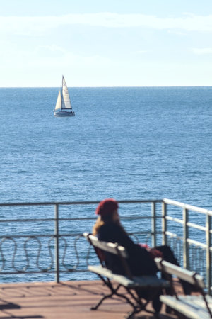 Sailing boat on the sea seen from a balcony with a woman out of focus sitting on a bench