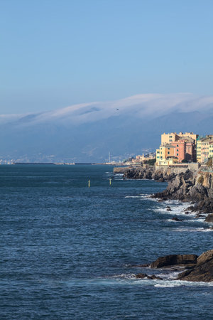 Town by the edge of a small cliff by the sea with a mountain ridge covered by clouds in the distanceの写真素材