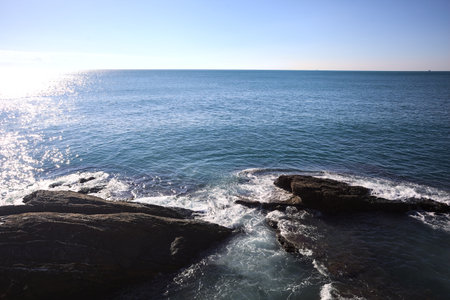 Waves of the sea crushing on the rocks of a cliff with sunlight cast on the water seen from aboveの写真素材