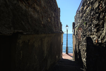 Narrow passage between stone walls that leads to a street lamps on a promenade by the sea on a sunny dayの写真素材
