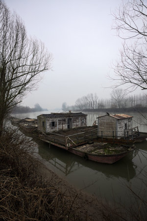 Riverbank below an embankment with a grove on it, with a moored houseboat, and the trees reflected in the water, and the opposite shore in the background seen on a cloudy and misty dayの写真素材