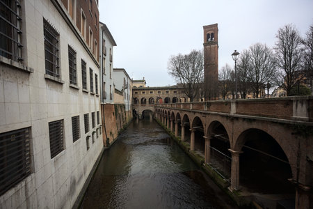 Stream of water passing next to a porchway by the shore, seen from above a panoramic balcony, with buildings and a tower in the background, on a cloudy day in an Italian townの写真素材