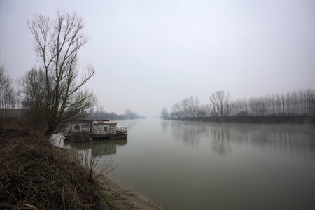 Riverbank below an embankment with a grove on it, with a moored houseboat, and the trees reflected in the water, and the opposite shore in the background seen on a cloudy and misty dayの写真素材