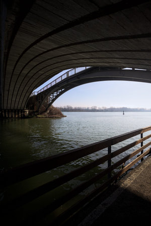 Counterlit arches of a bridge, and their beams, seen from a passage under it, by the water's edge,...