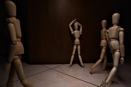 Wooden mannequin leaning against and facing a wall its arms on its face with other wooden mannequins...