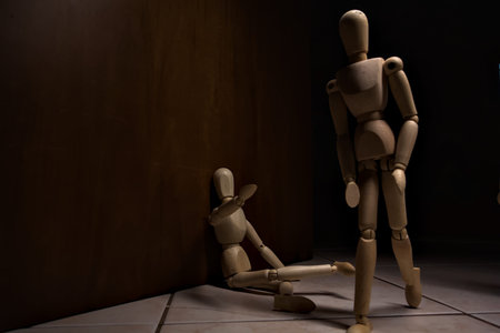 Wooden mannequin pretending to sit on a street and to lean on a wall raising his hand toward passing people in the act with other wooden mannequins in the act of passing by on a streetの写真素材