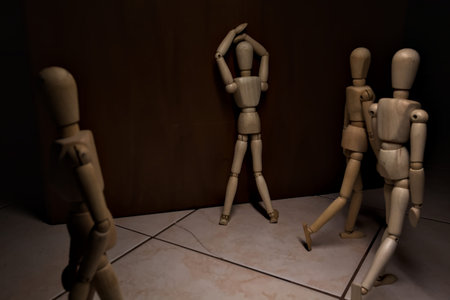 Wooden mannequin leaning against and facing a wall its arms on its face pretending to cry in despair with other wooden mannequins in the act of passing byの写真素材