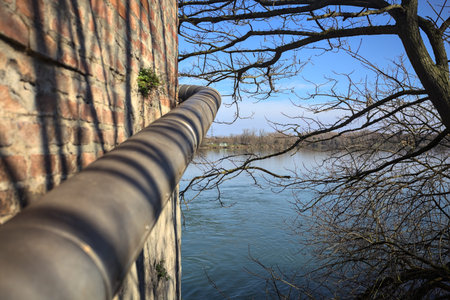 Lakeshore in the distance, framed by branches of a bare tree, a brick wall, and a pipe attached to...