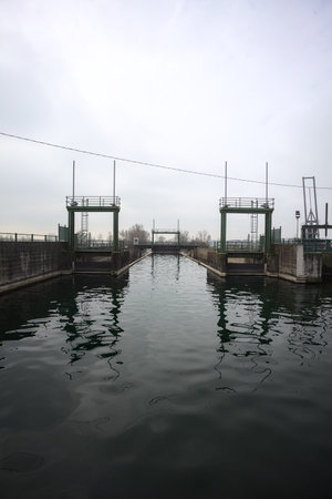 Sluices of a dam, seen from behind a diversionary channel above an embankment, with the sky cast on...